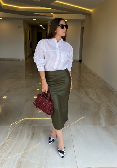 Camisa blanca de botones, falda lápiz de cuero verde oliva, bolso rojo y tacones de punta en patrón blanco y negro. Fondo de suelo de mármol.