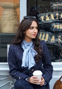 Tummansininen tweed-takki, jossa metallinhohtoisia yksityiskohtia, vaaleansininen silkkihuivi, pitää valkoista kahvikuppia, istuu koristeellisen ikkunan lähellä.