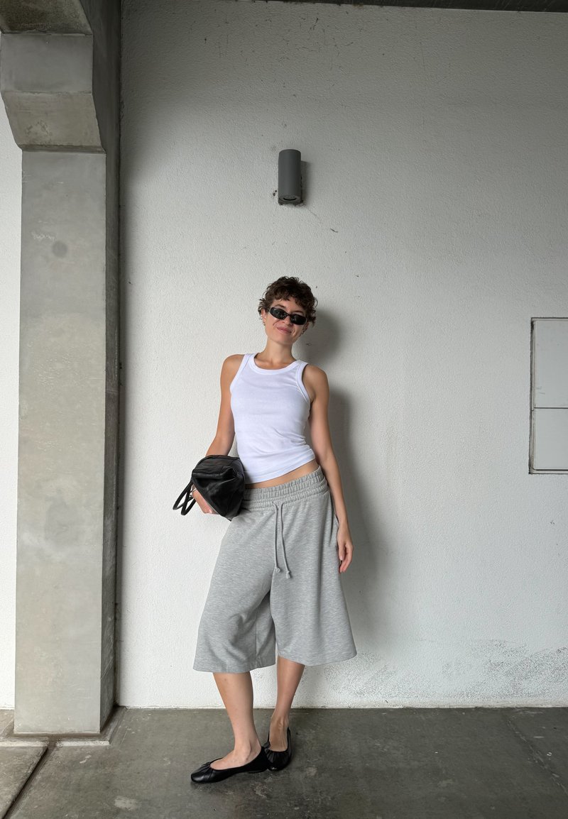 Camisola sem mangas branca, calções culotte cinzas, sapatos rasos pretos e uma bolsa preta. O cenário apresenta uma coluna de betão cinza e uma parede branca.