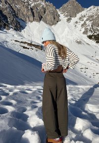Pull court à rayures gris clair, pantalon large kaki, paysage enneigé, bonnet bleu et bottes marron avec des accents texturés.