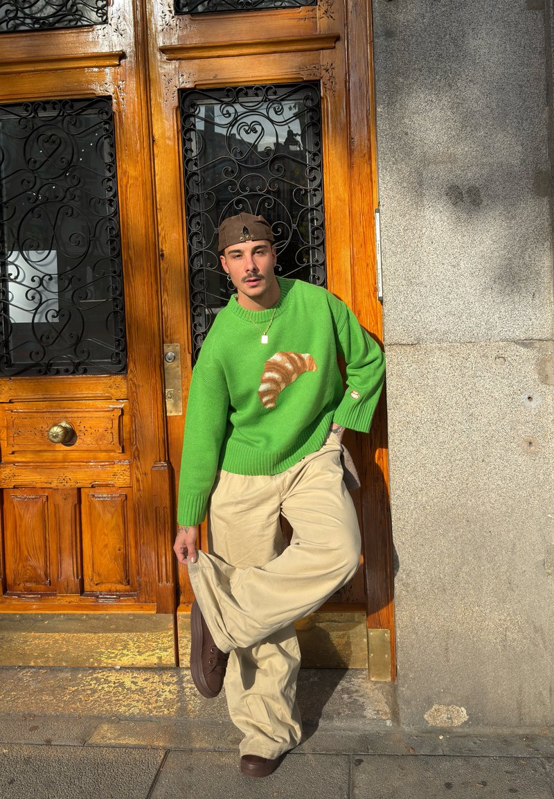 Homme portant un pull vert avec un motif de croissant, un pantalon beige et une casquette marron, appuyé contre une porte en bois ornée de ferronnerie sous la lumière du soleil.