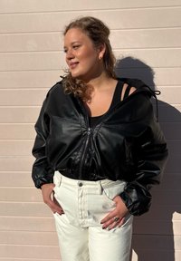 Svart faux läder kort jacka med dragkedja, elastiska ärmar och dragsko huva, i kombination med högmidjade vita jeans.