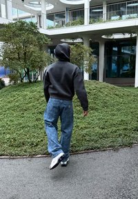 Pilkas džemperis su kapišonu, mėlynos plačios džinsinės kelnės su išblukusiu raštu ir balti sportiniai bateliai, einantys takeliu šalia žalumos ir modernios architektūros.