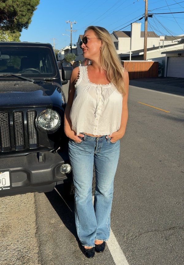 Femme portant un haut blanc sans manches, un jean bleu et des lunettes de soleil, debout à côté d'un SUV noir dans une rue ensoleillée.