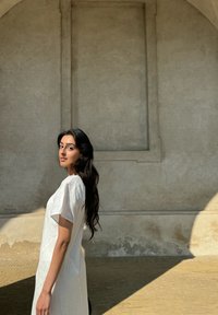 Fließendes weißes Kleid mit kurzen Ärmeln, strukturierter Stoff und lockere Passform, vor einer hellen Steinewand und Schatten.