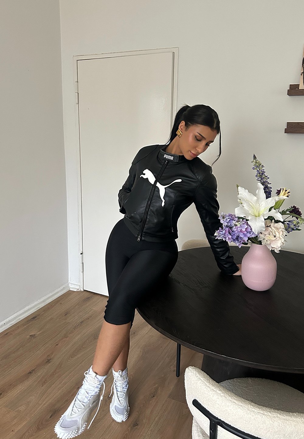 Blouson aviateur en cuir noir avec logo Puma blanc, shorts ajustés noirs, et baskets montantes grises. Table en bois courbée avec un vase rose et des fleurs.
