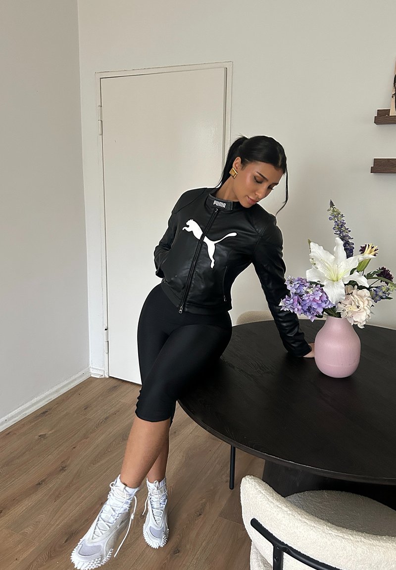Blouson aviateur en cuir noir avec logo Puma blanc, shorts ajustés noirs, et baskets montantes grises. Table en bois courbée avec un vase rose et des fleurs.