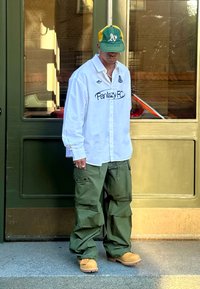 Fehér ing "Fantazy FC" felirattal, olajzöld katonai nadrág, sárga és zöld baseball sapka, és világosbarna lábbeli egy zöld ajtónál állva.