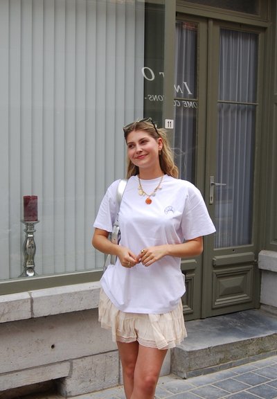 Camiseta blanca de algodón con mangas cortas, que presenta un pequeño gráfico. Combinada con pantalones cortos de tela arrugada de color beige claro, de pie frente a un edificio.