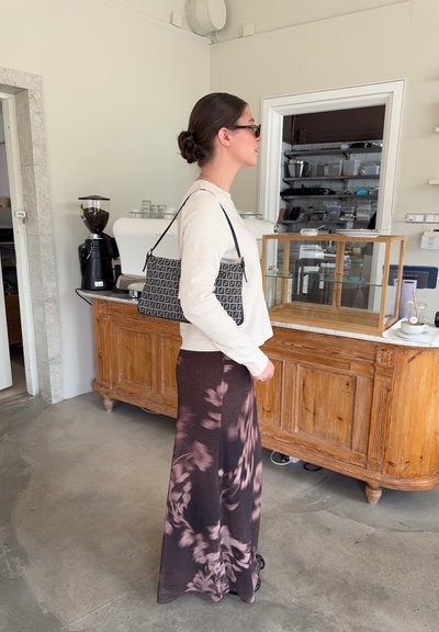 Bolso de mano de estampado negro y gris con el logo F; textura suave, forma rectangular. La modelo lleva una blusa color crema y una falda larga floral marrón.