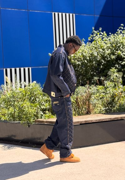 Chaqueta de mezclilla azul oscuro con patrón acolchado y detalles en amarillo, combinada con pantalones a juego y calzado en tono marrón claro, contra un fondo azul.