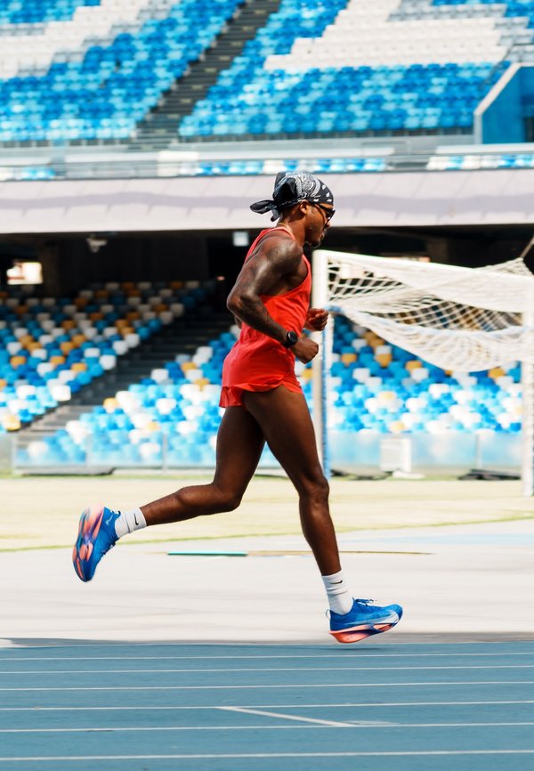Atleta vestido de rojo corriendo en una pista azul dentro de un estadio vacío con asientos multicolores y una portería de fútbol de fondo.