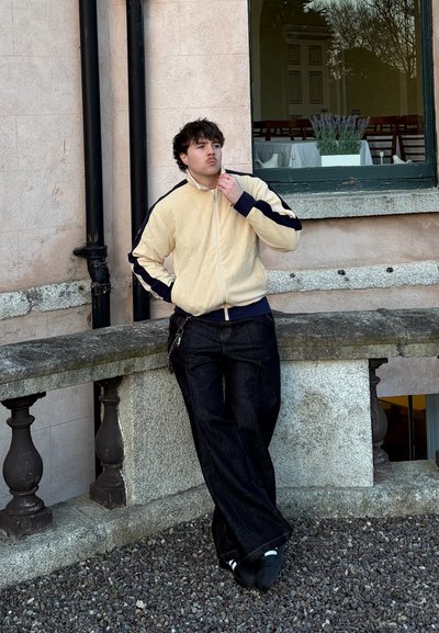 Joven con una chaqueta beige y azul marino apoyado en una barandilla de piedra fuera de un edificio con una ventana que muestra una mesa y una planta de lavanda.