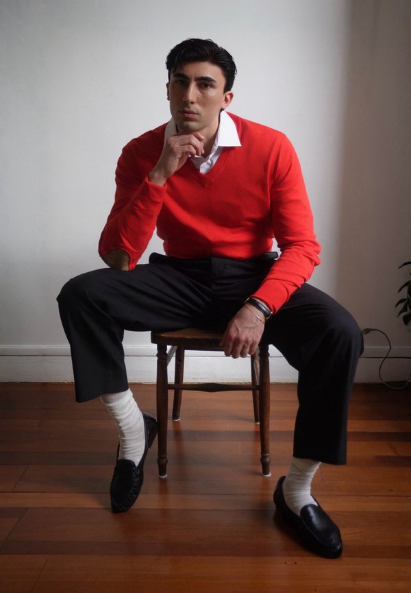 Mand med mørkt hår, iført rød sweater, hvid skjorte, sorte bukser, hvide sokker og sorte loafers, siddende på en træstol på trægulv.