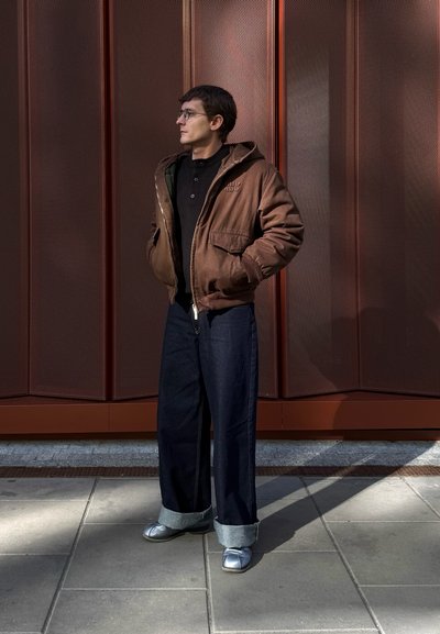 Chaqueta con capucha marrón sobre una camiseta negra, combinada con jeans oscuros de pierna ancha y zapatos metálicos plateados. El fondo presenta un panel de cobre texturizado.