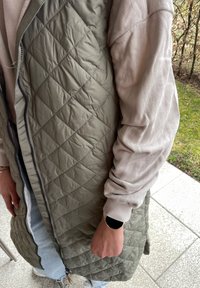 Κουβάρτινη ελιά πράσινη γιλέκο με φερμουάρ μπροστά, συνδυασμένη με ένα ανοιχτό ροζ μπλούζα με στρογγυλή λαιμόκοψη. Έχει ριμπ μανσέτες και χαλαρή εφαρμογή.