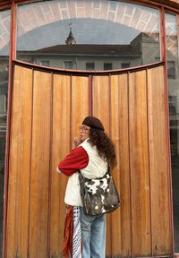 Persona ar cirtainiem matiem, valkājot krēmkrāsas šūtu vesti, sarkanu kreklu un brūnu bereti, stāv pie koka durvīm, turot rakstainu somu.