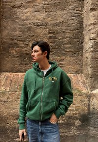 Green zip-up hoodie with a front pocket, featuring embroidered "BLU VIS NY." Paired with light blue jeans in front of a textured wall.