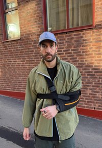 Olivgrön zip-up jacka med avslappnad passform, bärande en svart arm sling och klocka. Ljusblå keps; lager av svarta kläder synliga under.
