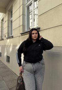 Μαύρο ριγωτό μπουφάν με φερμουάρ και λευκές ρίγες στα μανίκια, σε συνδυασμό με ψηλόμεσα ανοιχτό γκρι τζιν. Κρατάει μια καφέ τσάντα με μοτίβο.