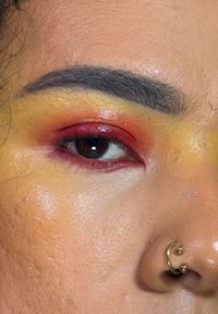 Gros plan sur un œil avec un fard à paupières vibrant orange, rouge et jaune. Présente un anneau de septum doré et une peau texturée.