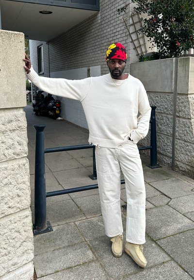 Un suéter extragrande de color beige claro y pantalones ligeros a juego, combinados con zapatos color marrón. Un gorro a rayas rojas y amarillas completa el look.