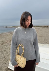 Γυναίκα με σκούρα μαλλιά μέχρι τους ώμους, ντυμένη με γκρι πουλόβερ και κρατώντας μια μπεζ τσάντα, στέκεται σε μια αμμώδη παραλία κάτω από συννεφιασμένο ουρανό.