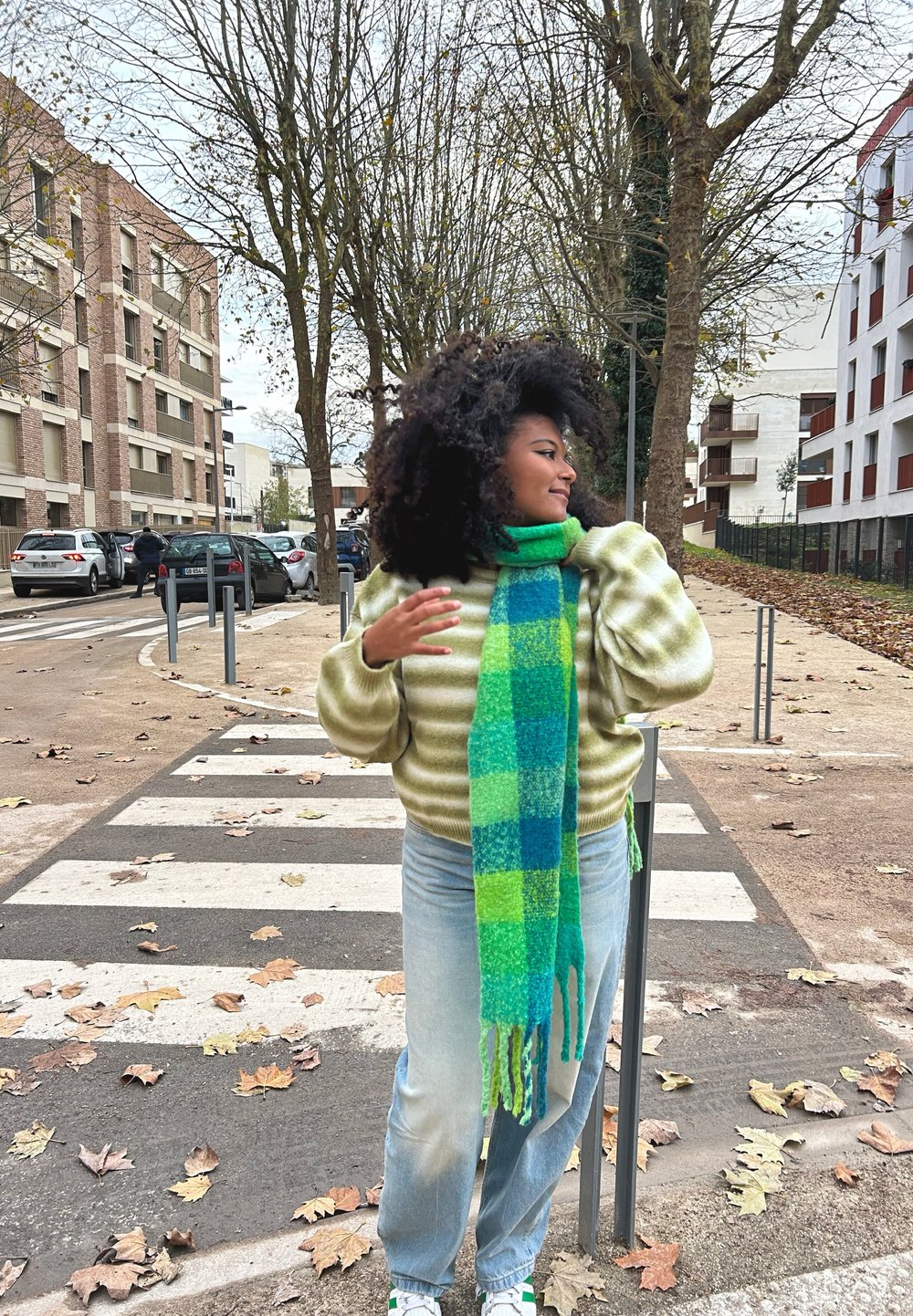 Πράσινο και μπλε καρετί μαντήλι, μαλακό πουλόβερ από μαλλί με ανοιχτό πράσινες ρίγες, ανοιχτού μπλε τζιν, στέκεται σε διάβαση πεζών περ�ιτριγυρισμένος από δέντρα.