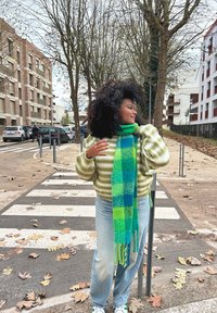 Πράσινο και μπλε καρετί μαντήλι, μαλακό πουλόβερ από μαλλί με ανοιχτό πράσινες ρίγες, ανοιχτού μπλε τζιν, στέκεται σε διάβαση πεζών περιτριγυρισμένος από δέντρα.