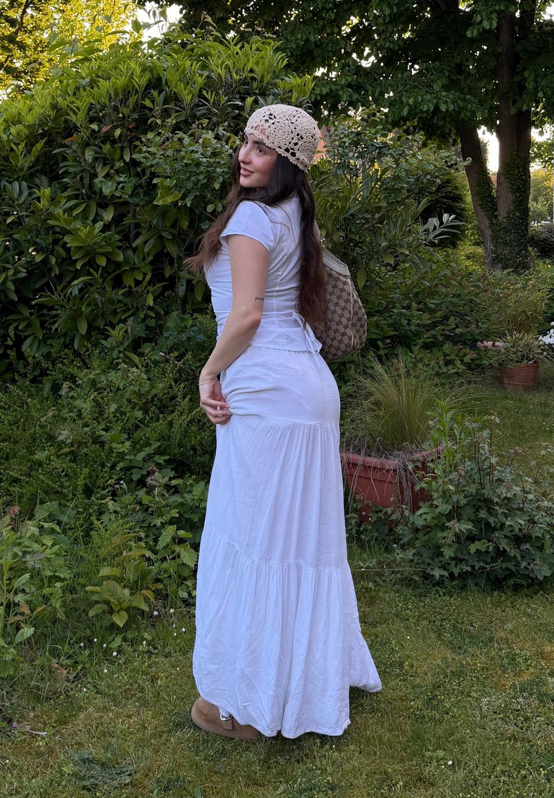 Mujer con vestido blanco y sombrero de ganchillo está en un jardín, mirando hacia atrás por encima del hombro, sosteniendo un bolso estampado.
