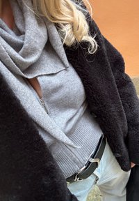 Personne portant un manteau noir en tissu pelucheux, un pull gris à fermeture éclair avec col châle, un jean en denim clair et une ceinture noire avec une boucle argentée.