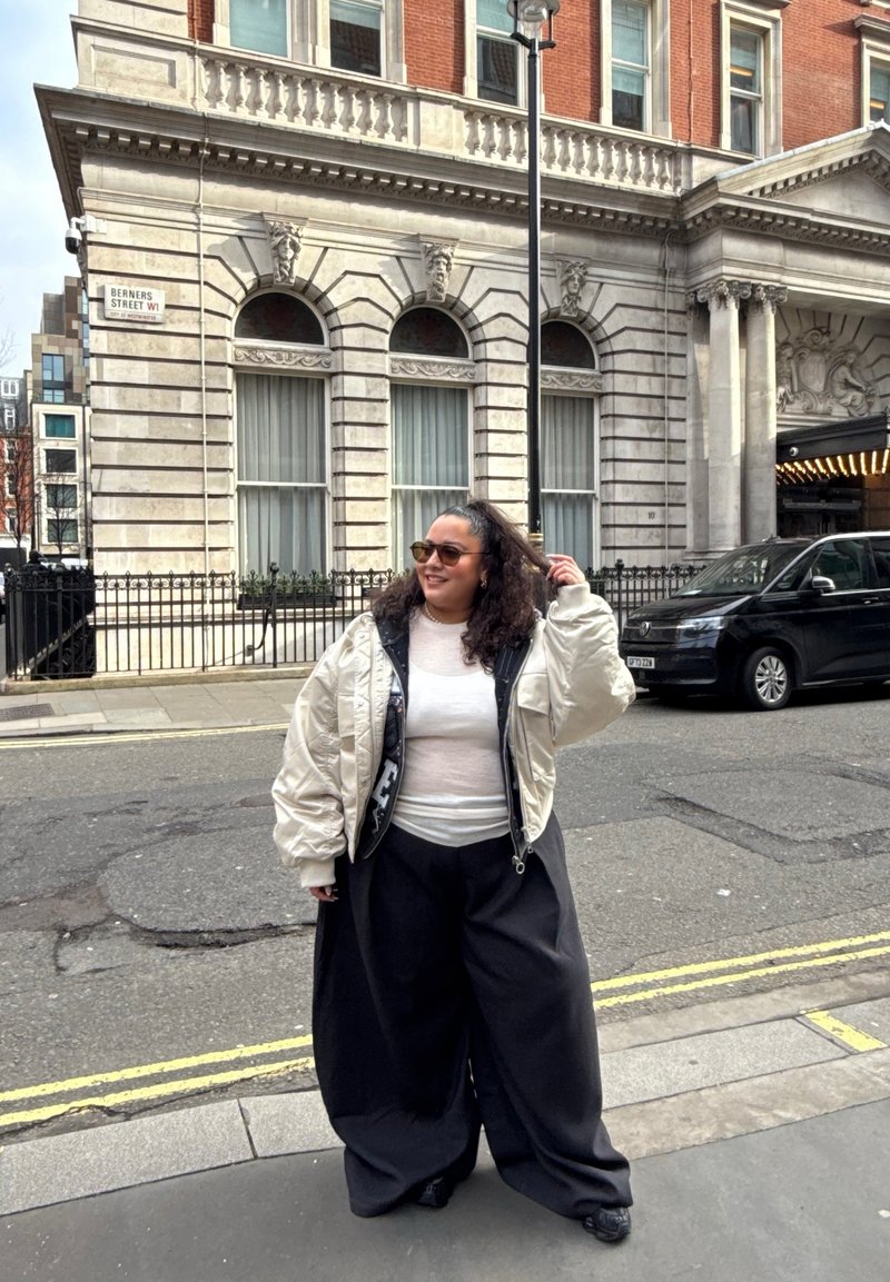 Femme aux cheveux bouclés portant des lunettes de soleil, une veste blanche, un haut blanc transparent et un pantalon large noir, debout à un coin de rue près de bâtiments historiques.