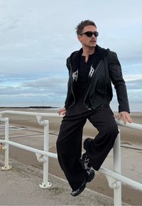 Muškarac u crnoj kožnoj jakni preko crne košulje s gumbima, obučen u široke karirane hlače i crne tenisice, stoji pokraj ograde.