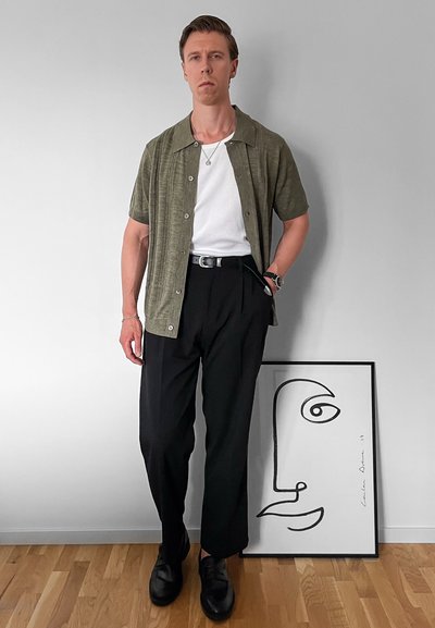 Camisa de botones de manga corta en un verde oliva claro sobre una camiseta blanca, combinada con pantalones negros y zapatos negros. Obra de arte abstracto en un marco cercano.