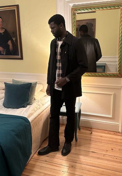 Hombre con abrigo negro y camisa de cuadros de pie junto a una cama con almohadas turquesas y manta en una habitación con paredes amarillas y suelo de madera.