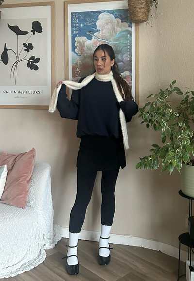 Suéter navy, mini falda negra, leggings negros, calcetines blancos y zapatos de charol negros. Presenta una bufanda blanca acogedora y vegetación de interior.