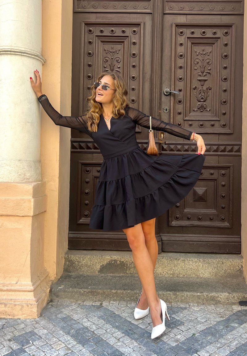 Μαύρο φόρεμα με φούστα σε στρώσεις, μακριά διαφανή μανίκια, V-ντεκολτέ και υφασμάτινη υφή. Λευκές μυτερές γόβες συμπληρώνουν το σύνολο, μπροστά σε ξύλινο φόντο.