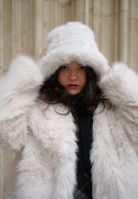 Plašč iz umetnega krzna v svetlo roza barvi z puhasto teksturo in velikim kapuco. Pod njim črni pulover. Temna podlaga z nevtralnim kamnom.