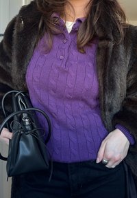 Pull en laine violet à torsades avec un col boutonné, superposé sous un manteau en fourrure sombre. Tient un petit sac noir avec un nœud décoratif.