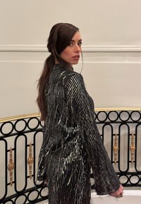 Femme aux longs cheveux bruns, vêtue d'une robe à paillettes rayée noire et argent, tournant la tête près d'une rampe ornée noire et dorée dans un intérieur au décor neutre.