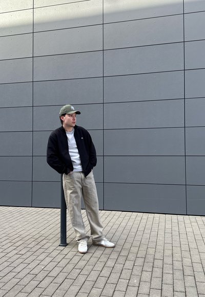 Joven con ropa informal y gorra se apoya en un poste negro cerca de una pared gris con azulejos sobre una superficie pavimentada al aire libre.