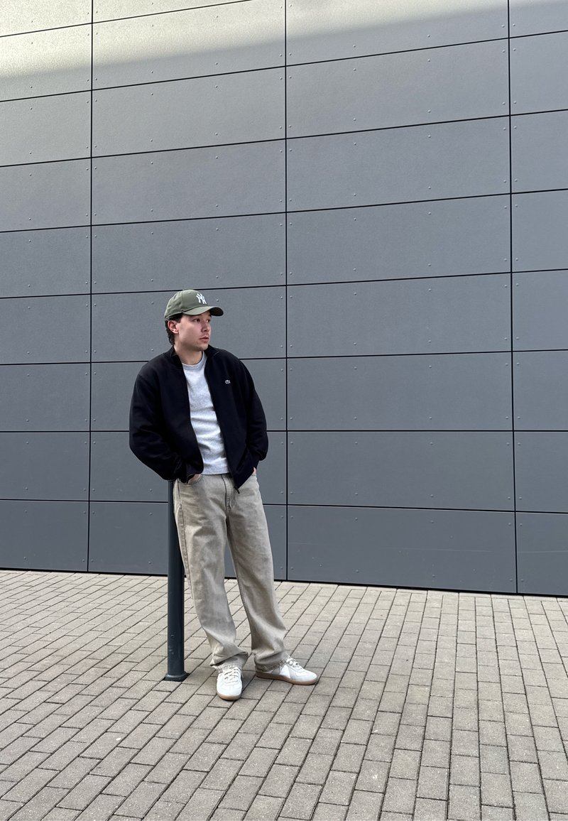 Jeune homme en vêtements décontractés et casquette appuyé contre un poteau noir près d’un mur carrelé gris sur une surface pavée en plein air.