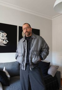 Mees prillide ja habemega, seljas hall jakk ja triibulised püksid, seisab toas diivani lähedal, seinal abstraktne kunst.