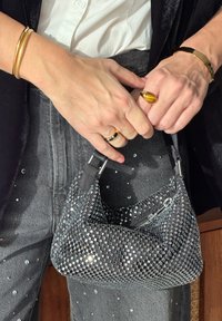 Srebrna i crna tkanina torba okruglog oblika, glatke teksture, s patentnim zatvaračem. Uparena s sivim hlačama koje imaju svjetlucave detalje.