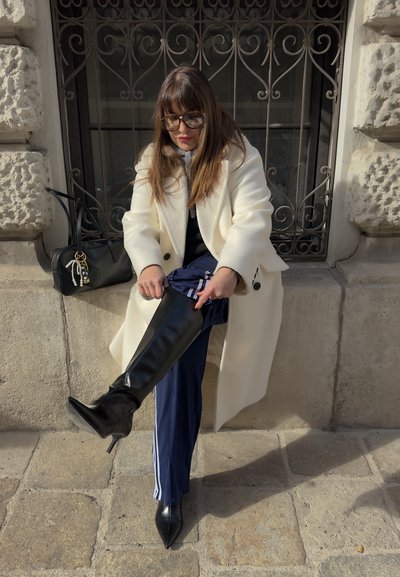 Botas negras por encima de la rodilla con punta afilada, combinadas con pantalones de chándal azul marino con rayas blancas. Un abrigo color crema complementa el look.