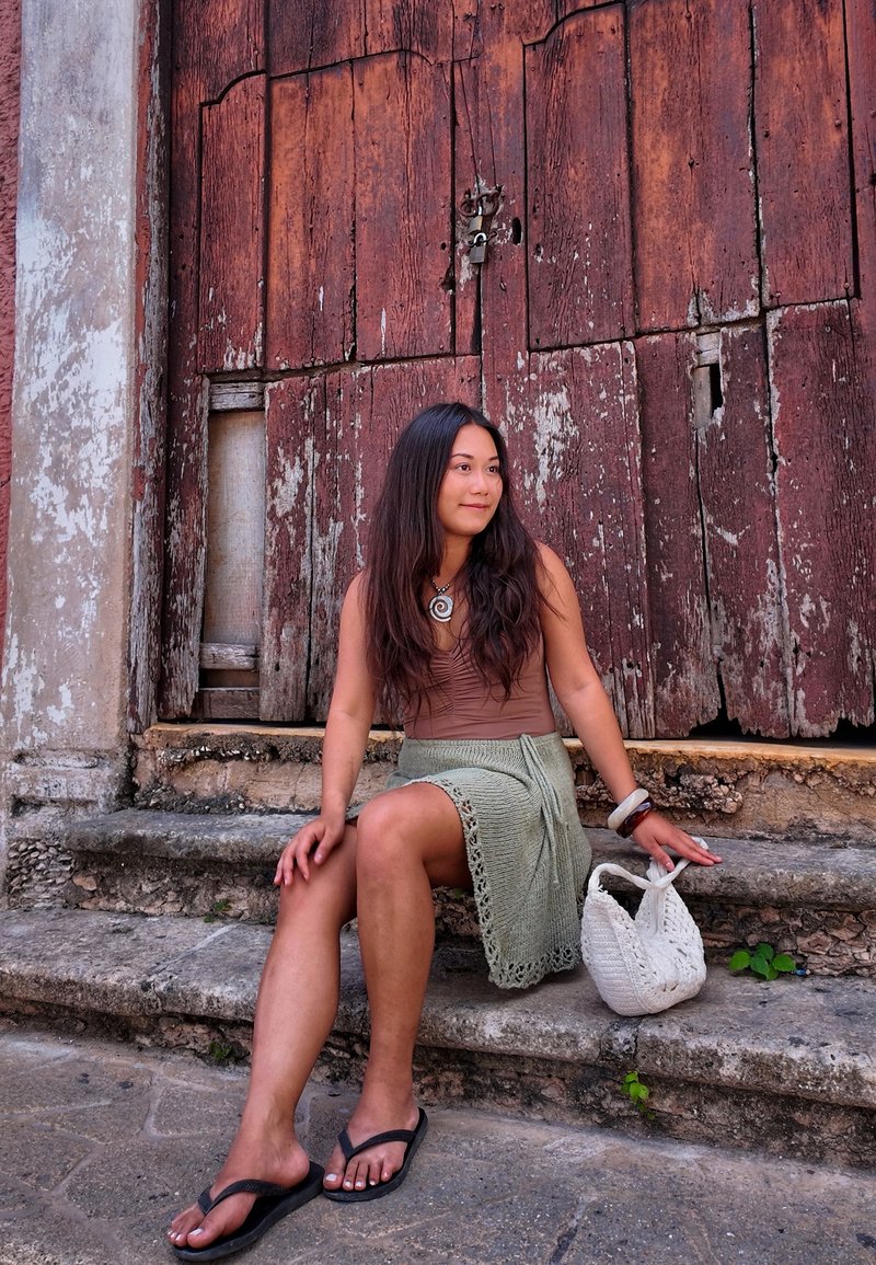 Mujer con cabello largo y oscuro, vestida con una blusa marrón y una falda verde, sentada en escalones de piedra junto a una bolsa blanca tejida, frente a una puerta de madera envejecida.