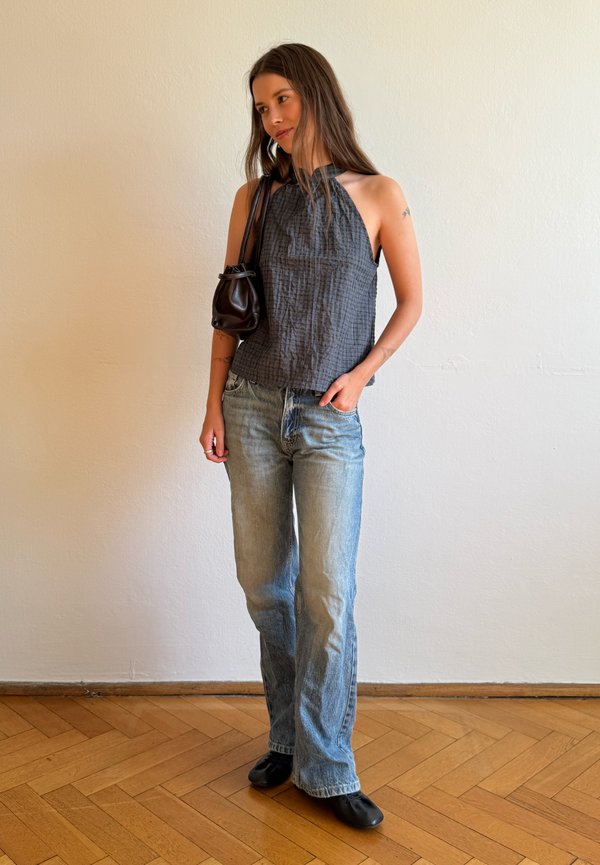 Giovane donna con top grigio scuro senza maniche, jeans azzurri, scarpe nere e piccola borsa nera a tracolla, in piedi su pavimento in legno.