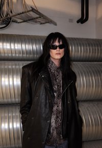 Veste en cuir noir portée sur une chemise à imprimé léopard. Le mannequin porte des lunettes de soleil noires. L'arrière-plan présente des conduits métalliques gris.