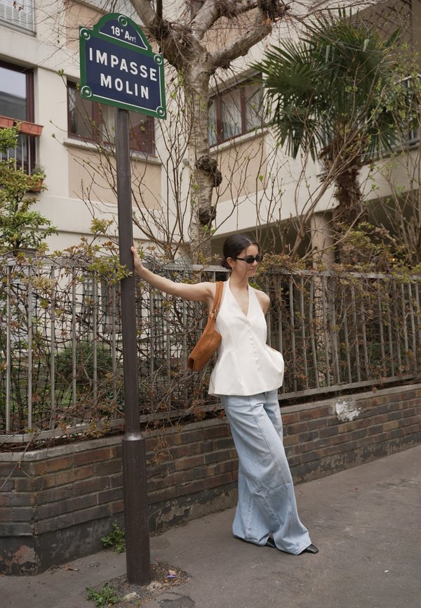 Woman wearing a white sleeveless top and wide-legged jeans leans on a street sign reading "Impasse Molin" near a brick wall and metal fence with trees.