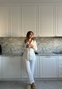 Set di pigiama bianco con bordi blu navy, in piedi in una cucina moderna con retrocucina in marmo, con in mano una tazza decorata, indossando pantofole beige.